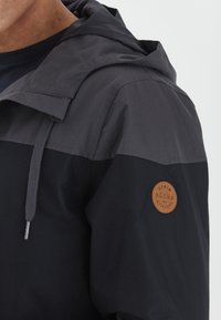 Kapa jakna s crnim i tamnosivim blok dizajnom, izrađena od glatkog materijala. Uključuje kožni logotip na rukavu.