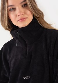 Czarny polarowy bluza z wysokim kołnierzem, pół zamkiem i wyhaftowanym na białym "COZY" na piersi. Miękka tekstura, luźny krój.