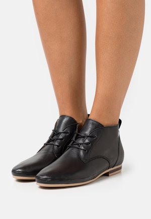 Czarne skórzane botki za kostkę z sznurówkami, noszone na gołych nogach, z płaską opaloną podeszwą i małym detalem na obcasie.