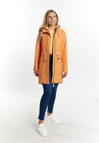 Veste de pluie orange vif avec une capuche, fermeture zippée, deux poches avant et cordons de serrage réglables ; portée sur une chemise rayée et un jean.