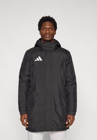 Manteau long noir matelassé avec capuche, présentant une fermeture éclair à l'avant, des poignets élastiques et un logo Adidas blanc sur le côté gauche de la poitrine. Texture lisse.