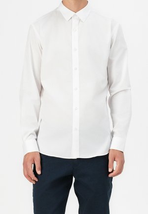Homme portant une chemise blanche unie à manches longues avec col et boutons, associée à un pantalon bleu marine foncé, debout devant un fond blanc.