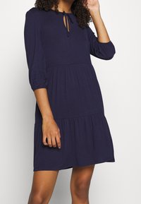 Kvinna som bär en mörkblå klänning med trekvartsärmar, ett volangkjol och en knytning vid nyckelhålet på halsen.