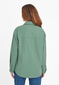 Green oversized shirt with a collar, long sleeves, and buttoned cuffs, made from a smooth fabric, featuring a back yoke seam.