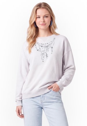 Giovane donna con lunghi capelli biondi indossa una felpa grigio chiaro con delicata stampa di catena e fiocco, jeans azzurri e orecchini a cerchio blu.