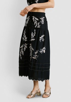 Femme portant une jupe midi noire avec des broderies florales blanches et un ourlet festonné, associée à des sandales à talons blocs à brides métalliques.