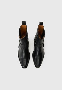 Botas de tobillo de cuero negro con punteras afiladas, que cuentan con una correa de hebilla lateral, parte superior texturizada y acabado suave. Interior en cuero marrón.