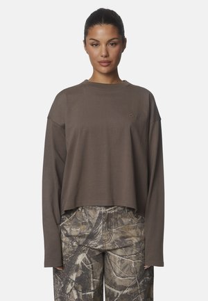 Donna con capelli scuri raccolti in uno chignon indossa una maglia marrone larga a maniche lunghe e pantaloni con fantasia mimetica, in piedi davanti a uno sfondo semplice.