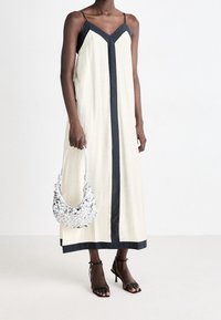 Ljusbeige maxiklänning med svarta detaljer och axelband, med en V-ringad design. Bärs tillsammans med en silverfärgad paljettväska och svarta klacksandaler.