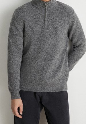 Man draagt een grijze halflange trui met een hoge kraag en donkere broek, staand tegen een effen lichtgekleurde muur.