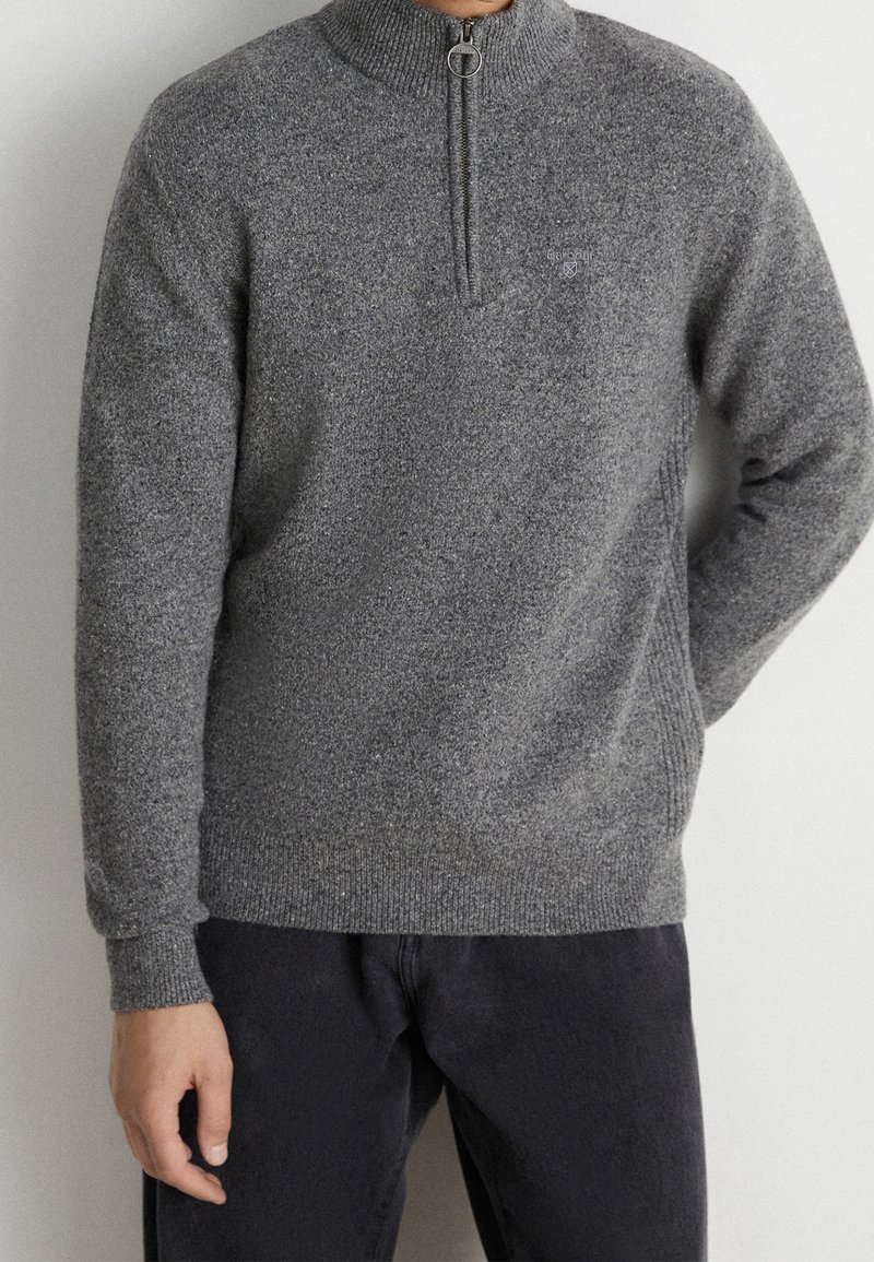 Homme portant un pull gris à demi-fermeture éclair avec un col montant et un pantalon foncé, debout contre un mur clair uni.