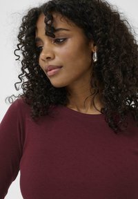 Maroon sweater with a round neckline and small textured patterns, paired with silver hoop earrings and curly dark hair.