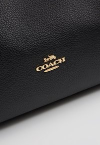 Surface en cuir texturé noir avec un petit logo doré Coach représentant une calèche tirée par des chevaux au-dessus du nom de la marque.