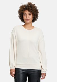 Helles beiges Pullover-Sweatshirt mit langen, ballonförmigen Ärmeln und Rundhalsausschnitt; weiche Textur und nahtloses Vorderdesign; getragen mit schwarzen Hosen.