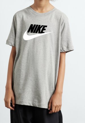 Barn iført en grå Nike t-shirt med sort Nike tekst og hvid swoosh-logo foran, står mod en ensfarvet baggrund.
