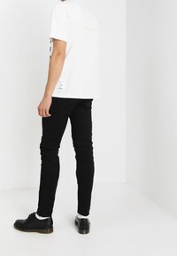 Vit t-shirt med kort ärm, svart text och en gul logotyp på baksidan, kombinerad med svarta slim fit-jeans och svarta skor.
