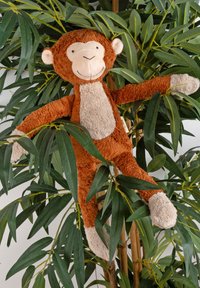 Brown plush monkey toy with a light beige face and belly, extending limbs, nestled among green leaves in a planter.