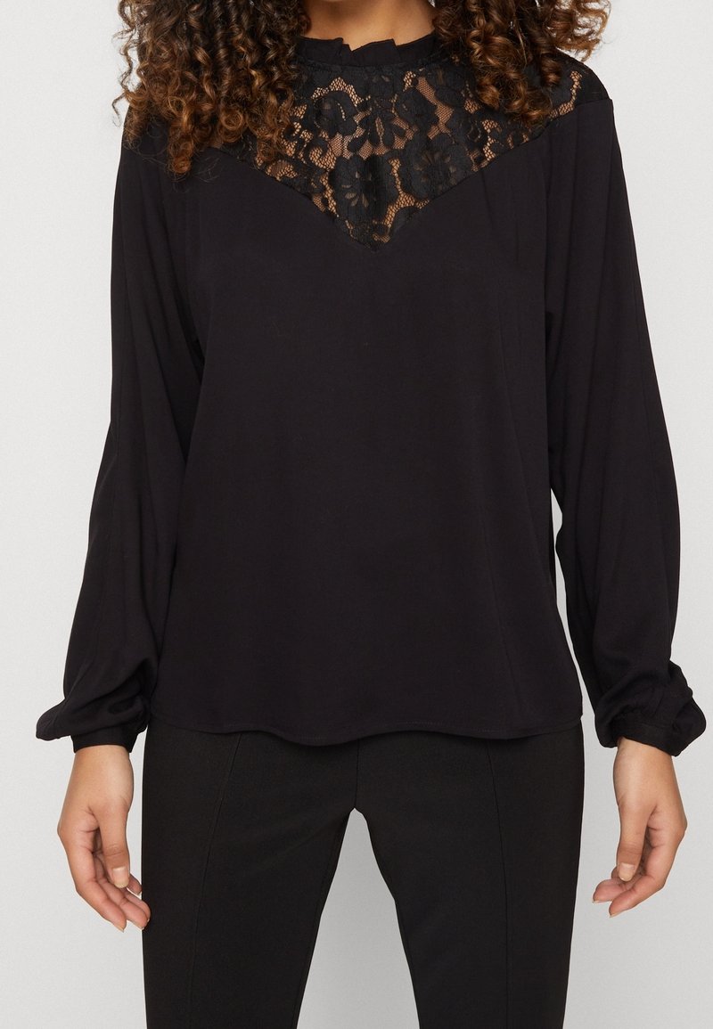Blouse noire à manches longues avec un design de yoke en dentelle, dotée d'un col rond et de poignets légèrement froncés. Finition en tissu lisse.