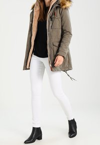 Femme portant un parka vert olive doublé de fourrure, un haut noir, un pantalon blanc et des bottines noires, debout devant un fond uni.
