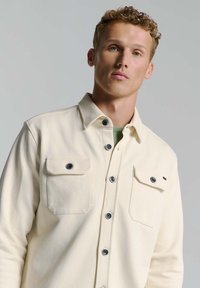 Crème knoopshirt met een kraag, twee borstzakken en donkerblauwe knopen. Gemaakt van gestructureerde stof, heeft het een gestructureerde pasvorm.