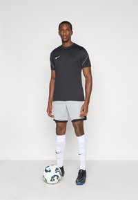 Μαύρη κοντομάνικη φανέλα Nike και γκρίζες βερμούδες, με λευκές και μαύρες λεπτομέρειες, συνδυασμένες με μαύρα παπούτσια ποδοσφαίρου και λευκές κάλτσες. Πλησίον υπάρχει μία μπάλα ποδοσφαίρου.