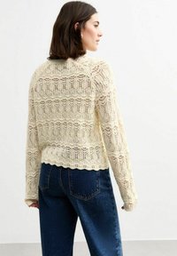 Pull en dentelle crème à manches longues, arborant des motifs ouverts complexes et un ourlet festonné, associé à un jean en denim bleu foncé.