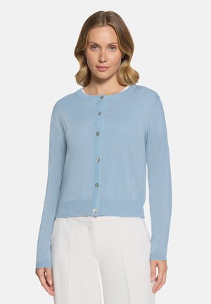 Woman with wavy blonde hair wearing a light blue buttoned cardigan and white pants, standing against a plain background.