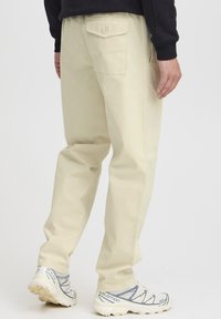 Pantaloni bej din bumbac, cu croială lejeră, prevăzuți cu un buzunar în spate cu închidere cu nasture, asortați cu pantofi sport albi cu accente gri.