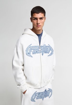 Giovane uomo che indossa una felpa bianca con zip e pantaloni abbinati con scritta ricamata blu "faster" su entrambi i capi.