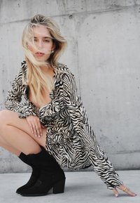 Sukienka koszulowa w zebrę z długim rękawem, czarne zamszowe botki za kostkę na słupku, na tle neutralnego betonowego tła.