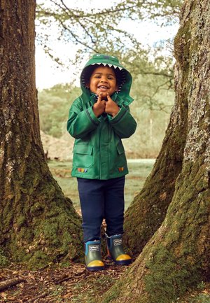 Copil zâmbitor cu pelerină verde de ploaie cu dinozaur și cizme colorate stă între trunchiuri de copaci acoperite cu mușchi, într-un cadru forestier.