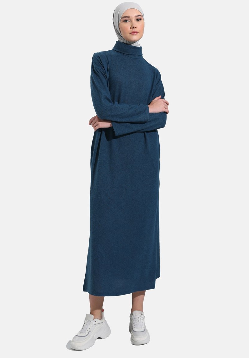 Abito a collo alto lungo di colore blu scuro, con maniche lunghe, realizzato in tessuto morbido; indossato con sneakers bianche. Texture liscia, design minimale.