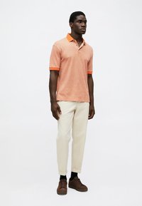 Orange polo shirt with contrasting collar and sleeves, featuring a textured pattern. Worn with cream trousers and brown sneakers.