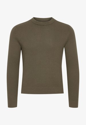 Mann i olivgrønn strikkegenser, mørkeblå jeans og svarte sneakers, står med en hånd i lommen og ser til siden.