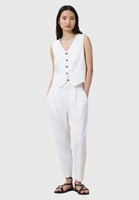 Weißes Top mit V-Ausschnitt und schwarzen Knöpfen, kombiniert mit eng geschnittenen weißen Hosen. Das Model trägt flache schwarze Sandalen. Glattes Material, minimalistisches Design.