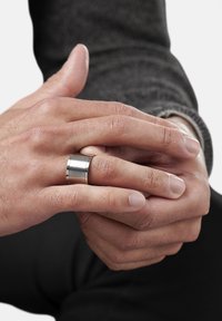 Metallring med borstad yta, med en slät, bred band. Händer visar naturlig hudstruktur; bakgrunden är enkel och ljus.