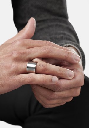 Metallring med børstefinish, som har en glatt, bred bånd. Hender viser naturlig hudtekstur; bakgrunnen er enkel og lys.