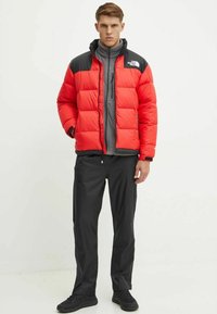 Giacca imbottita rossa con spalle nere, fodera interna grigia e tasca frontale con cerniera; indossata sopra uno strato intermedio grigio e pantaloni neri a vestibilità slim.