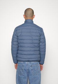 Giacca a vento in blu spento con un design trapuntato e strutturato. Caratterizzata da un colletto alto e polsini elasticizzati. Indossato con jeans in denim blu.