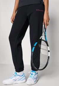 Čierne tepláky s manžetami na členkoch, s ružovým logom "BJÖRN BORG". Skombinované s bielymi a modrými športovými topánkami a tenisovou raketou.