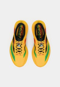 Chaussures de course jaune vif avec des accents verts et rouges, intérieur noir et semelles intérieures à motif graphique, vues de dessus sur fond blanc.