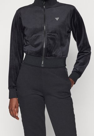 Chaqueta negra de tela suave con cremallera, bolsillos frontales y cintura elástica, combinada con pantalones negros a juego, llevada por una persona de pie.