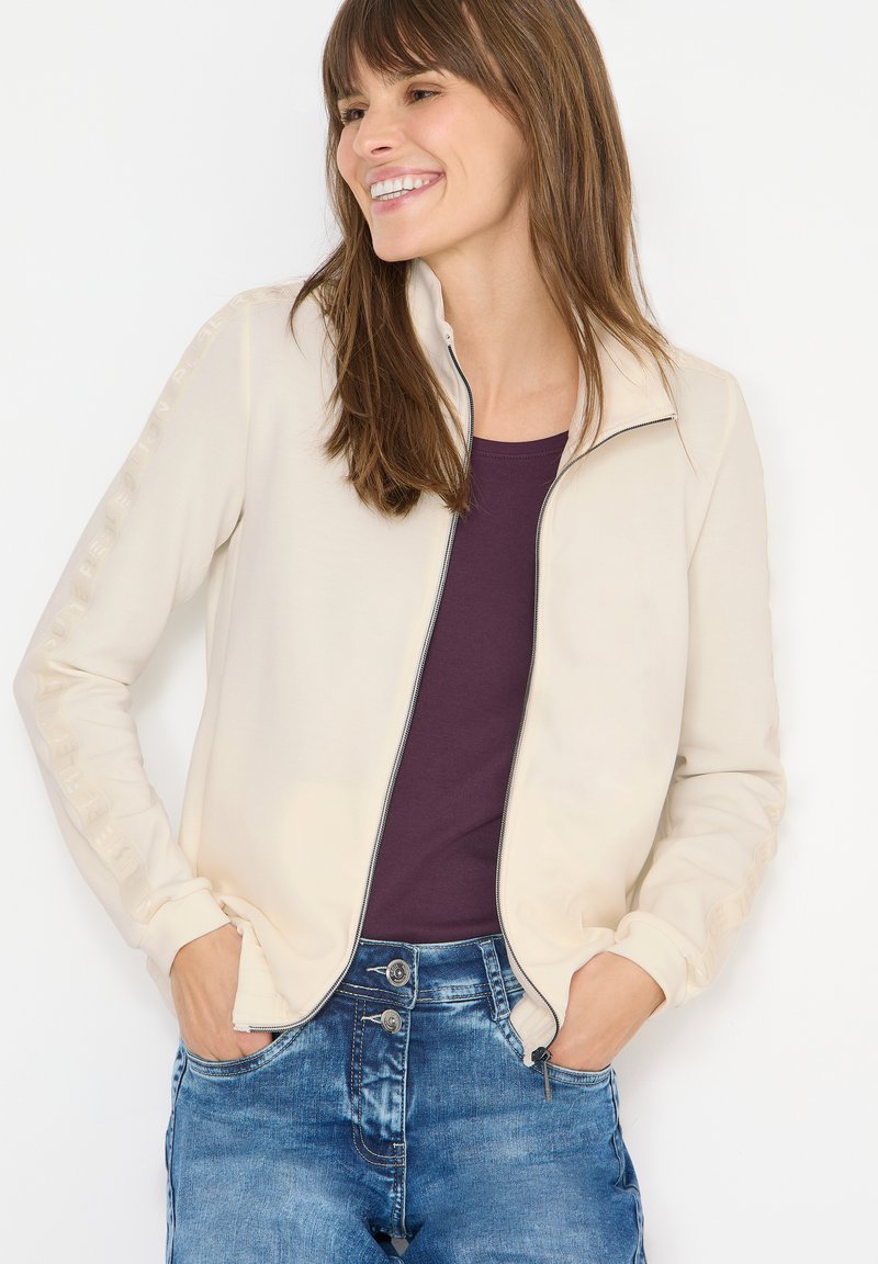 Chaqueta cremosa de color beige con cremallera y cuello alto, con una textura suave y detalles decorativos en las mangas. Combinada con una blusa morada.