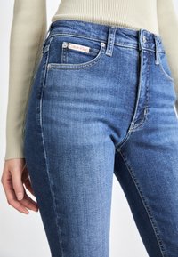 Jeans in denim blu con design a vita alta, stile a cinque tasche e etichetta con logo Calvin Klein. Texture liscia con leggere sfumature.