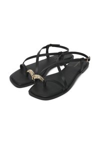 Sandales plates noires avec des fines lanières, détail en forme de croissant doré au niveau des orteils et boucles réglables à la cheville sur un fond blanc.