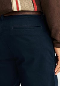 Sötétkék chino nadrág sima textúrával, hátsó zsebbel, és derékon látható barna bőr övvel. Finom varrási részlet.