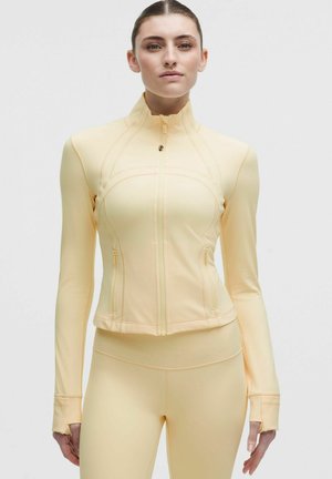 Donna che indossa una giacca aderente giallo pallido con cerniera e fori per i pollici, abbinata a leggings a vita alta, in piedi davanti a uno sfondo uniforme.