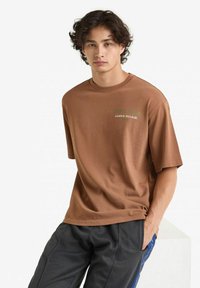 T-shirt in cotone marrone con vestibilità rilassata, caratterizzato da testo del logo verde sul petto. Il modello lo abbina a pantaloni grigio scuro.