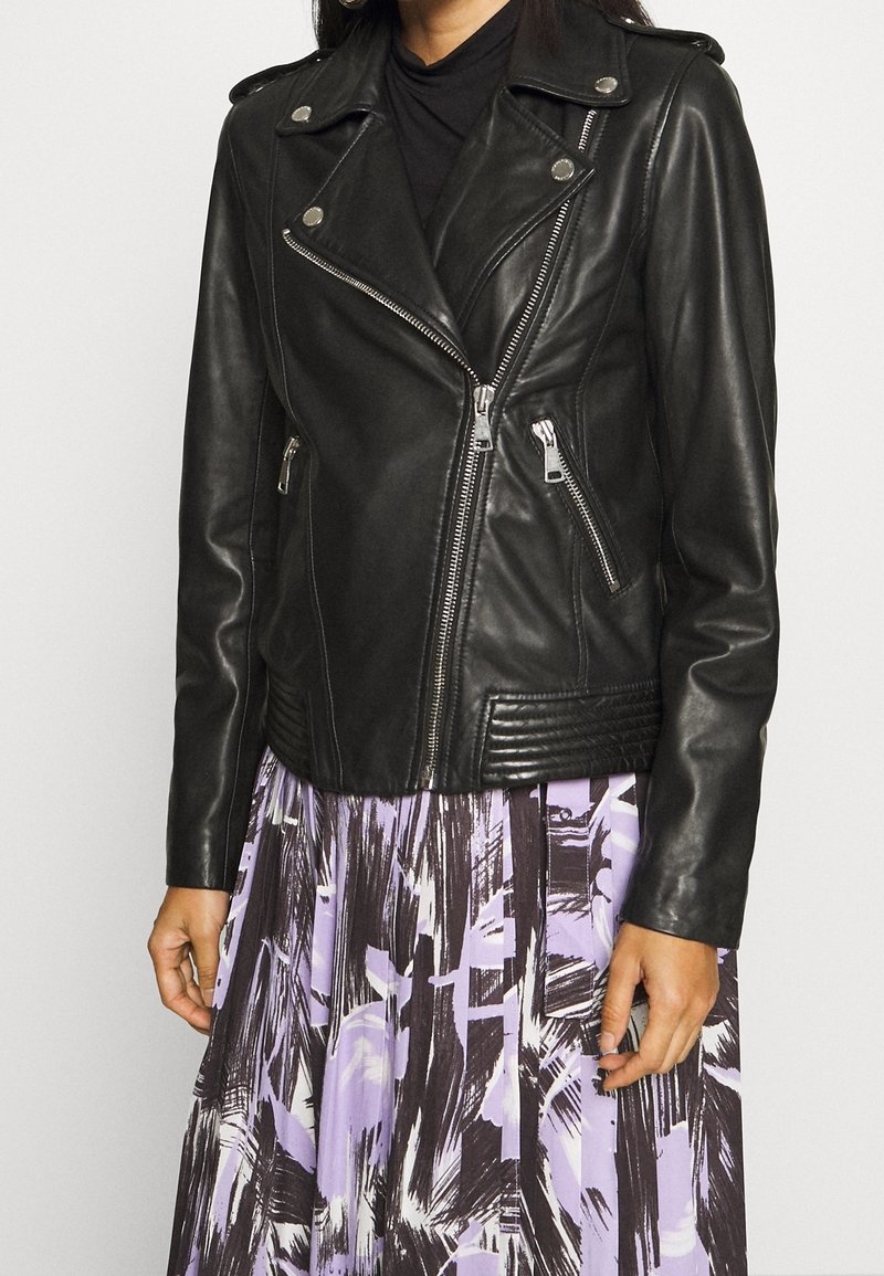 Schwarze Leder-Bikerjacke mit silbernen Beschlägen, diagonalen Reißverschlüssen und geripptem Muster am Saum, kombiniert mit einem farbenfrohen, gemusterten Rock.