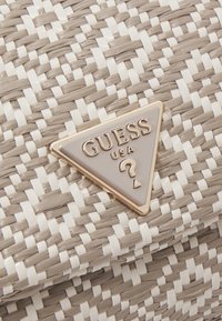 Sac en toile tissé gris et blanc avec une surface texturée ; présente un accent logo Guess triangulaire doré et gris.
